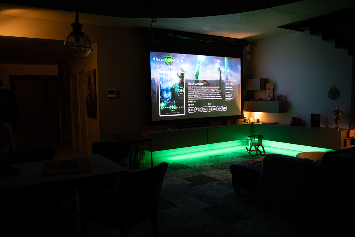 salon éclairé de couleur d'ambiance vert sous la télévision avec canapé, tables, chaises, vue sur la porte d'entrée